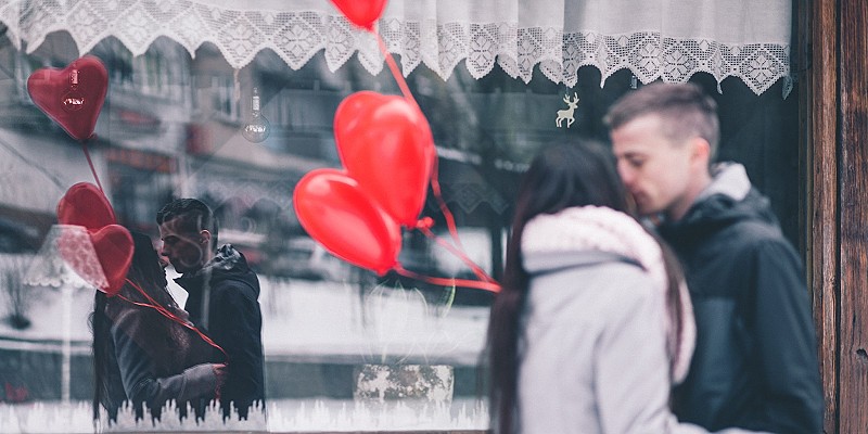 man and a woman kiss on the street with two red heart ballons