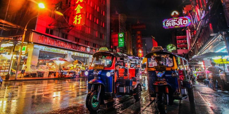 Two tuk-tuks wait on the street at night in Bangkok under the light of colourful neon signs