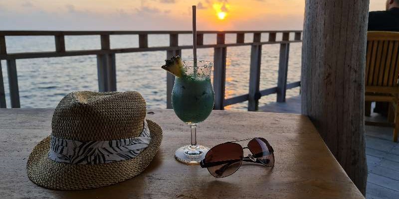 A hat, sunglasses and a drink on a table in front of an ocean view and sunset scene