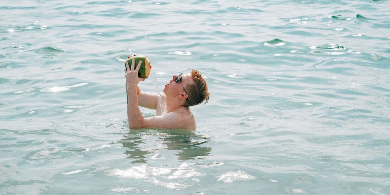 A man in standing in shoulder height water trying to drink from a coconut